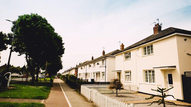 A row of lightly-coloured rendered houses. The one in the foreground has a white picket fence, with others having hedges along the front. There is a green space to the front of the properties.