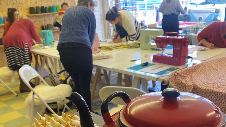 A group of people stood around a table with sewing machines on it with a pot of tea and a cake in the foreground