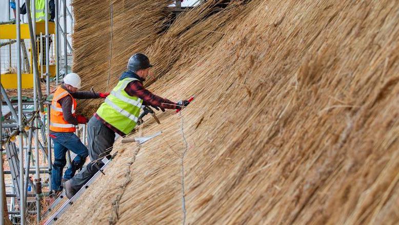 Workers step on a ladder to re-thatch the Tithe Barn