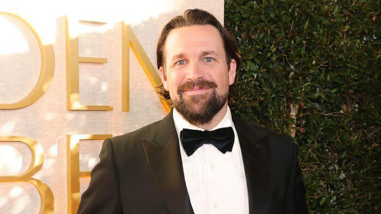 Trevor Matthews is looking at the camera, he is stood in front of a sign which says Golden Globes in gold font, but you can only read the 'D E N' of golden and the 'B E' of globes. He is wearing a black tuxedo with a black bow tie and a white shirt. He is smiling at the camera and his dark hair has a middle parting, he has a moustache and beard in the style of a goatee.