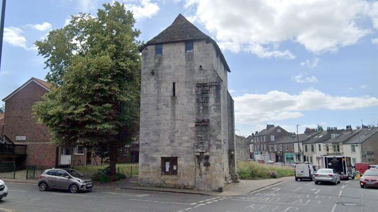 Fishergate Postern Tower stands in front of a tree and houses at a road junction with cars queuing in traffic nearby