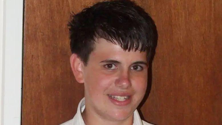 Jimmy Mizen smiling. It is a close up photo. He has dark hair and appears to be standing in front of an oak coloured door