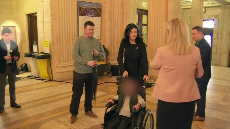 A photo from the event at Stormont showing Michael McMonagle standing to the left of the screen looking down at his phone, he is wearing trousers, shirt and a jacket. Michelle O'Neill stands to the right of the screen with her back to the camera, wearing a pink jacket. In the centre of the image is a child in a wheelchair with their face blurred and their parents standing behind him.