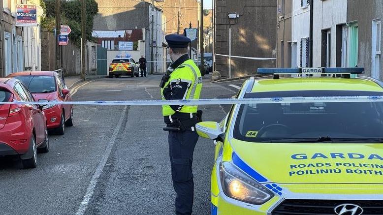 A street in New Ross is cordoned off after a fatal assault on a young girl.  A uniformed garda (Irish police) office stands guard at the cordon tape besisde a yellow and blue patrol car.  Another yellow and red emergency vehicle is in the background at the other end of the cordon. 