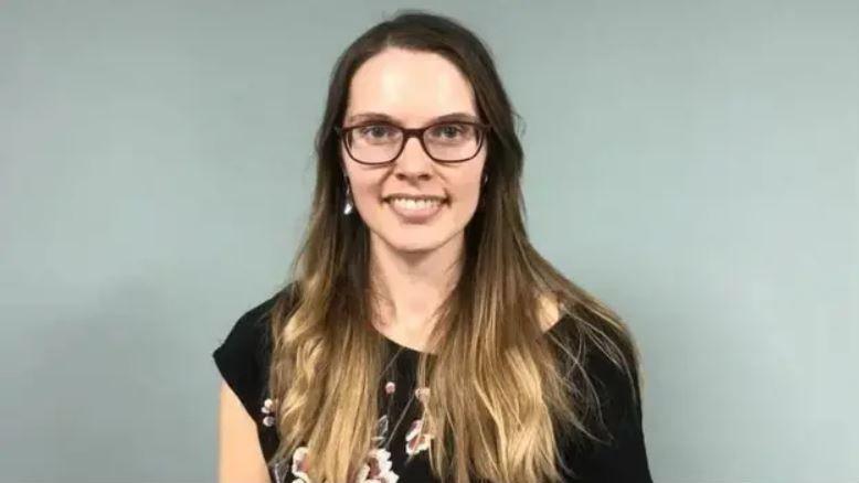 Joney Faragher looking straight ahead smiling. She has long brown hair and is wearing glasses and a black top.