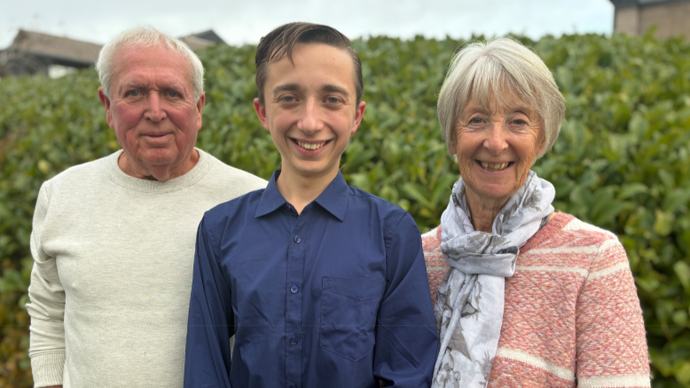 Adrian, Daniel and Jill in a midshot in front of a tall hedge all smiling to camera.