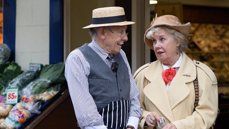 An older man stands next to a woman, both looking cheerful. They appear to be in conversation, and both are dressed in 1940s style clothing and hats. They're standing in front of a greengrocers.