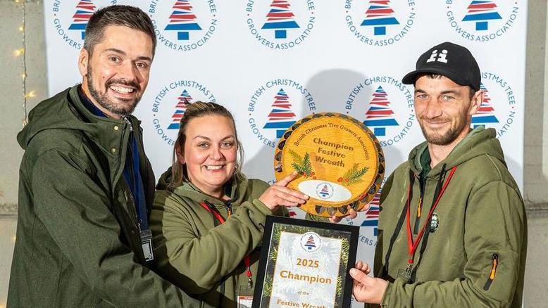 Two men and a woman in green jackets stand in front of a backdrop with logos for the British Christmas Tree Growers Association, featuring red and blue Christmas tree illustrations. They are holding awards and celebrating. The circular wooden plaque reads "Champion Festive Wreath" and the framed black certificate reads "2025 Winner". There is a grey brick wall in the background, with a wooden table to the right and a string of yellow fairy lights hanging down on the wall.