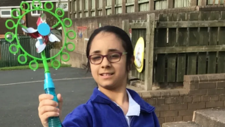 Shay is in her blue school uniform in what could be her school's playground holding a large, green plastic toy towards the camera. She has dark hair tied back and is wearing glasses.

