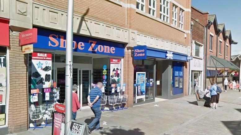 Google Maps image of the former Shoe Zone. Two people are walking past the building while nearby two more people are sitting on a bench talking to a third person.