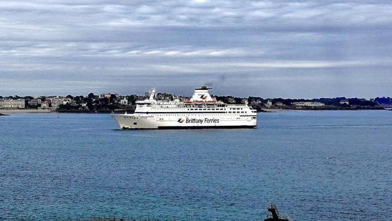 A white Brittany Ferries sails into a harbour with a coastal town in the background.