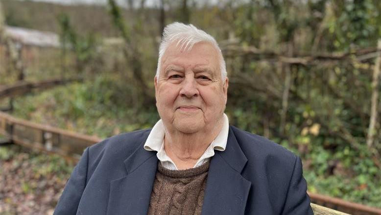 An elderly man with white hair, smiling as he looks at the camera. He is wearing a white shirt with brown knitted jumper and dark blue blazer. He is stood in front of a blurred brown fence with green hedges and trees in the background