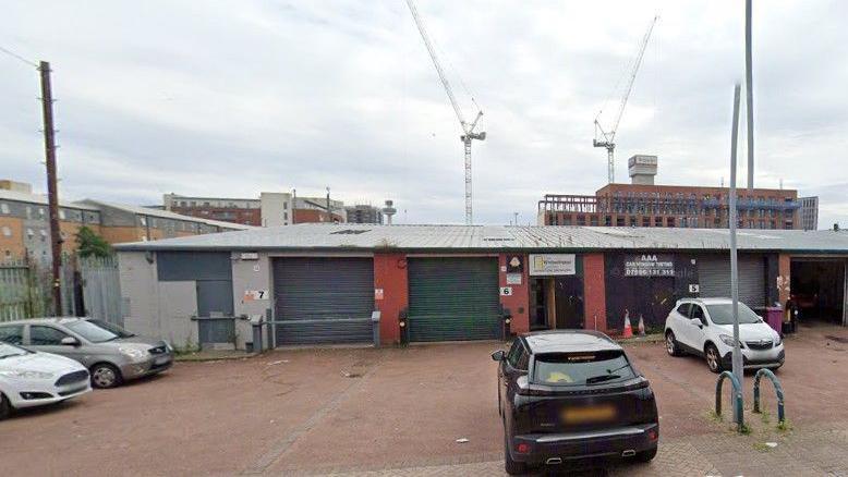 A warehouse with shutters down has a sign saying 'Whitechapel Centre'. There is a car park at the front with a number of cars parked. Large buildings and cranes can be seen in the distance