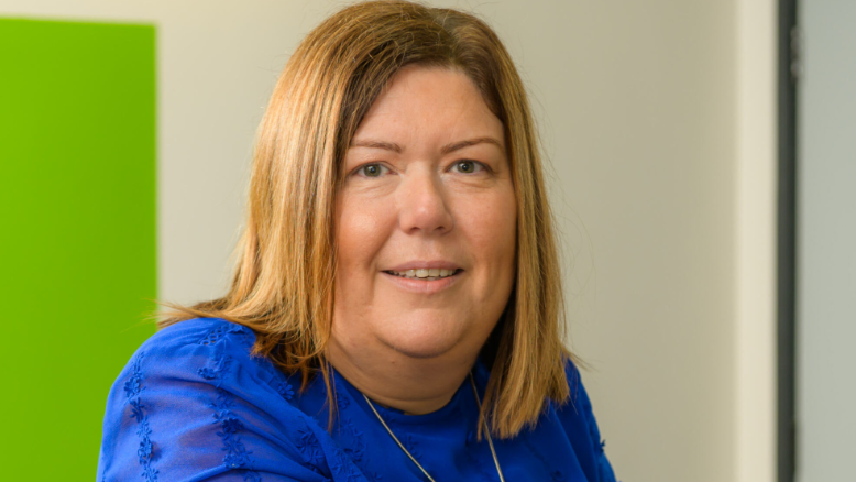 Michelle is standing in front of a white and green wall wearing a blue top and gold necklace chain. She has blond shoulder length hair and brown eyes. She is smiling at the camera. 