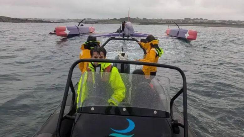 A black boat being driven by a man with short black hair in high visibility clothing. There are two others behind him looking out to a vessel that has capsized.