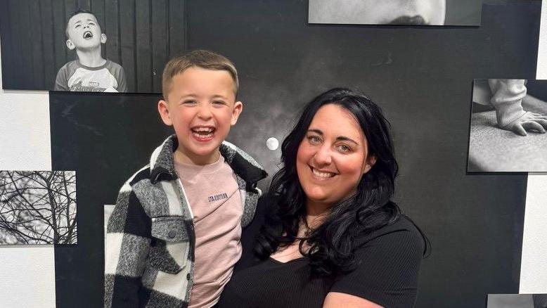 Bo Willow Harris, in a black top and leopard print skirt is holding her son Hendrix. They are both smiling at the camera while standing in front of black and white photographs attached on a white wall as part of the exhibition. Hendrix has short dark blonde hair and is wearing a black and white jacket, pink t-shirt, and a black trousers. His mother has long wavy black hair.