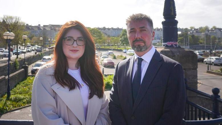 A man and a woman stand side-by-side near the Queen's Parade area of Bangor.
The woman has long reddish hair and is wearing glasses, a light coloured white T-shirt and overcoat. The man next to her is wearing a dark suit and blue tie. He has light stubble and short hair.