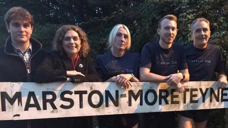 Five people line up behind a sign saying "Marston Moreteyne". Three are wearing running shorts and blue t-shirts with "CAPTAIN TOM" in white writing. The other two are wearing black fleeces and one is Hannah Ingram-Moore. There are trees in the background.