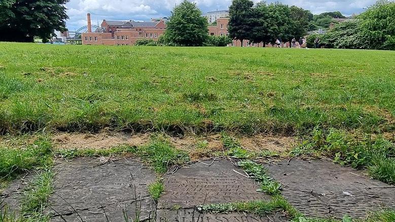 A path made out of a headstone surrounded by grass. Houses and the Tyne Bridge can be seen in the background.