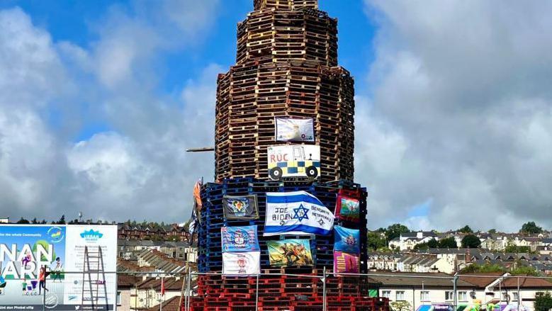 A bonfire in Derry's bogside made from pallets and covered in flags including unionist items and the flag of Israel
