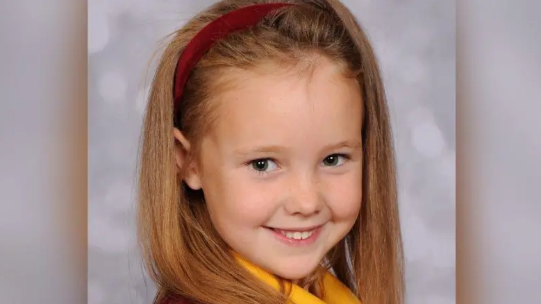 A headshot of Elsie Dot Stancombe. She had long brown hair, tied back with a red headband, and is smiling.
