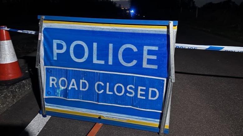 A police closed sign on the guided busway between Histon and Cambridge. The sign is blue with police road closed written on it in white letters. Behind it is blue and white police tape and on the right an orange and white road cone. In the distance can be seen a bright light from working emergency services. 