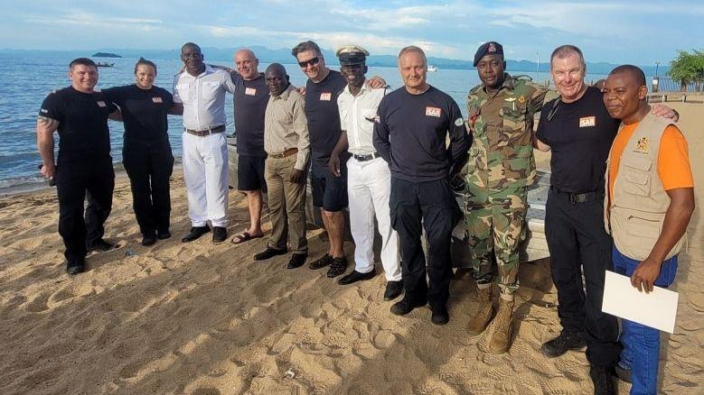 A line of 11 people including six firefighters, wearing their black uniform, stood alongside five Malawian delegates from the Defence Force, police, Port Authority and Department of Fisheries, all on the beach smiling at the camera.  