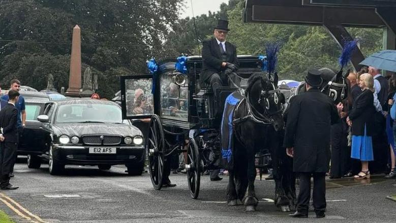 A horse-drawn carriage brings Jay Slater's coffin at the head of a cortege of cars to the chapel, with attendees lined up 