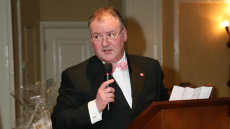 A man with grey hair and a white shirt and dark jacket and pink bow tie
