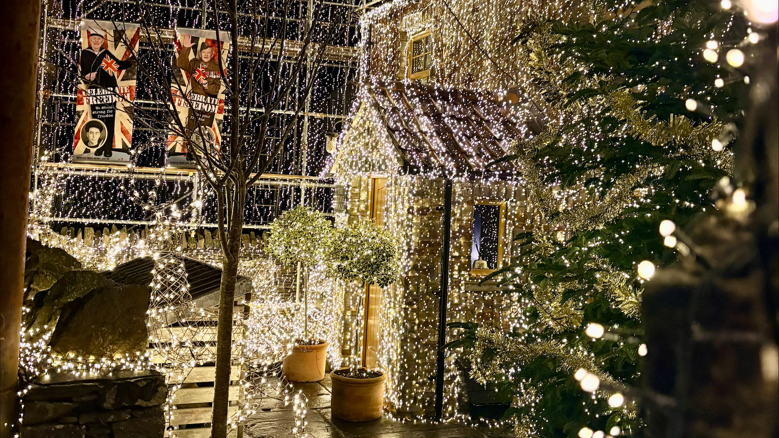 The front door and porch of a home, with lights draped all over. The rest of the building can be seen behind, also covered in white string lights. There is a large Christmas tree to the right of the front door.