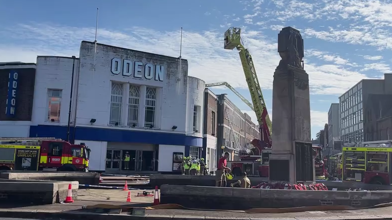 The front of an Odeon cinema, surrounded by fire engines and firefighters. A tall ladder which is higher than the building is parked next to the cinema, and is being used to extinguish a rooftop fire.