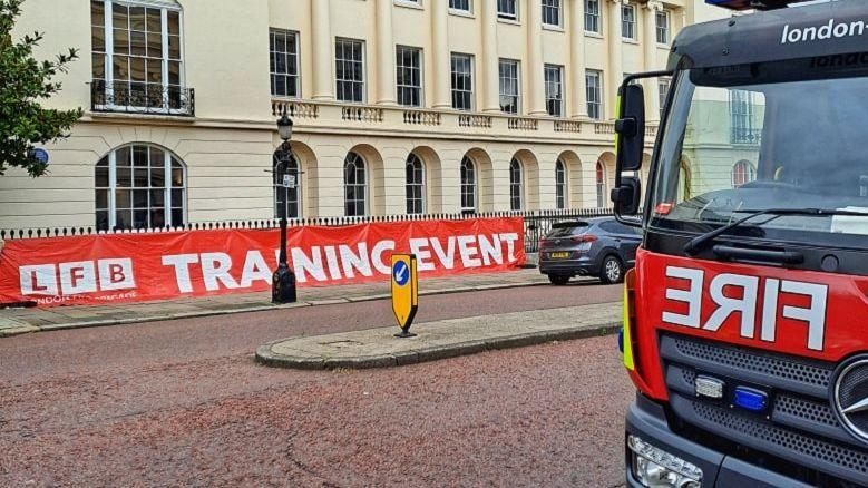 Part of a fire engine in shot, where the words FIRE can be seen. In the background is a cream building with pillars, with the words LFB TRANING EVENT on a banner along a gate. 