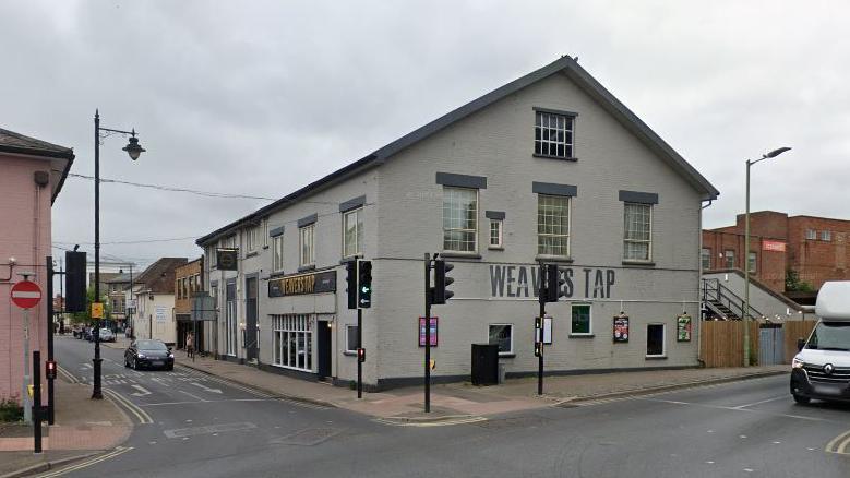 The Weavers Tap pub - a three-storey building on the corner of a town centre crossroads. It has a light grey painted exterior with dark grey lintels about the first and second floor windows. 