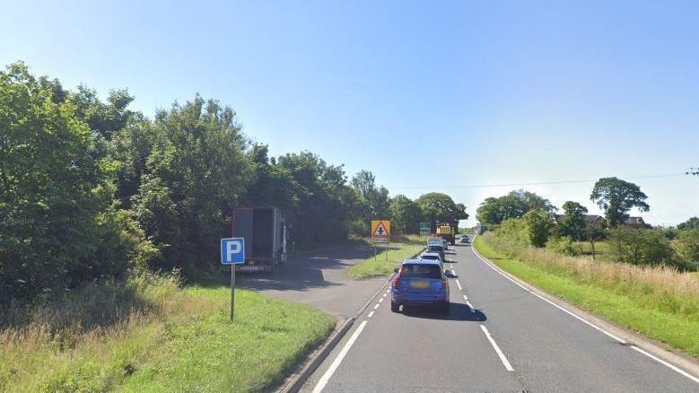 A layby on the A7. A lorry is parked in the layby and has its rear doors open. Several car are travelling on the single carriage road.