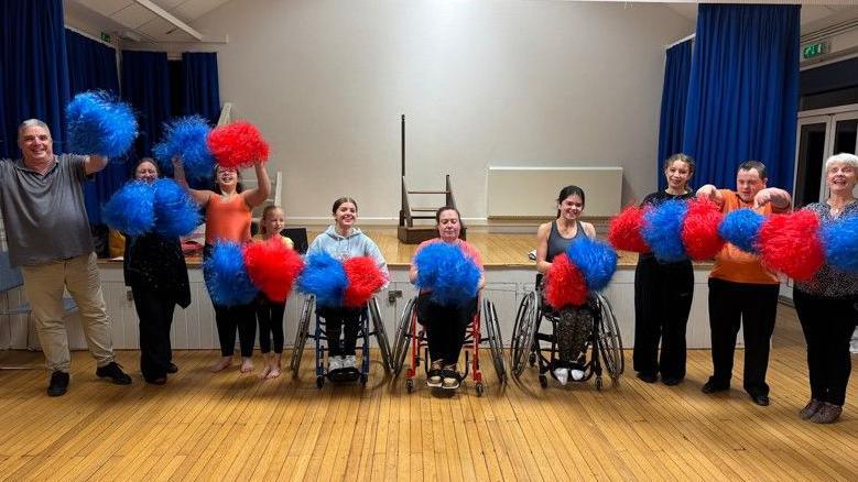 A mix of parents, children and their coach all stand or sit in a row, holding blue and red pom-poms. They are all smiling and cheering