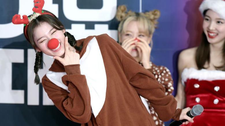 Sana from TWICE pictured at a photocall in Seoul in 2019. She is wearing a brown and white costume and reindeer antlers and a red nose. She is leaning to one side smiling at the camera. There are other people in Santa outfits in the background.

