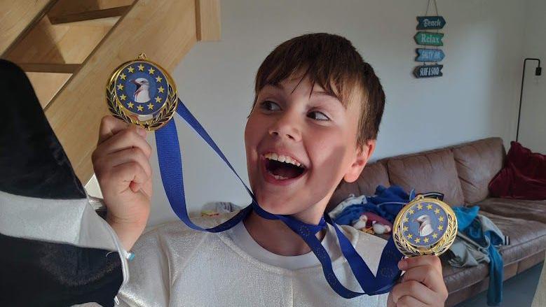 A boy smiles as he holds up two blue and gold medals with seagulls on them