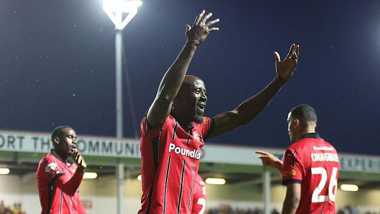 Albert Adomah has his arms up in the air celebrating a recent goal, wearing his Walsall home red and black kit