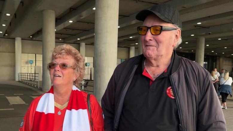 A woman with tinted glasses and a red and white scarf standing next to a man with yellow glasses on and a black Whitstable Town FC polo shirt.