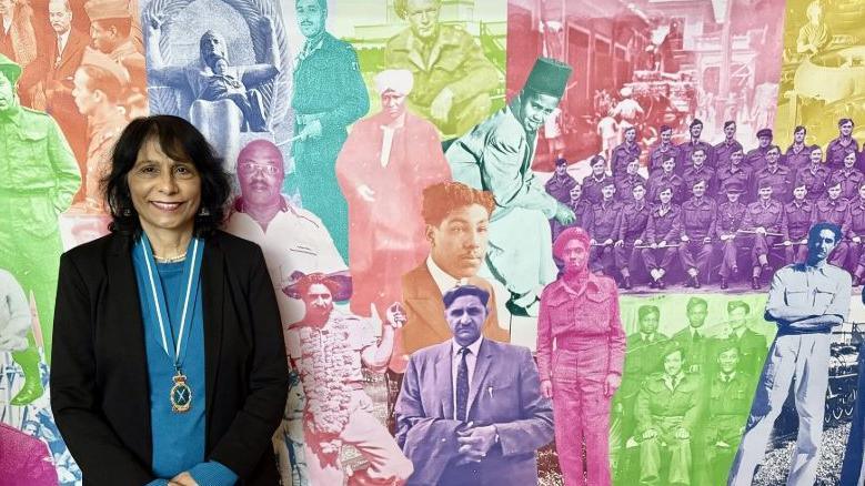 Ms Woolf stands in front of a montage of faces making up the photo banner. Each person's picture is a different colour. Ms Woolf wears a black jacket with a blue jumper underneath. She has a medal around her neck with a blue and white tie and a gold medal.