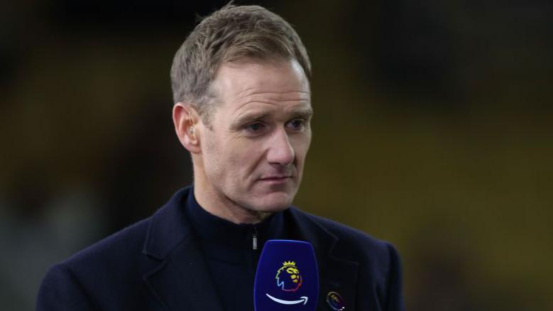 Dan Walker wearing a navy jacket and black zipped up top, holding a Premier League navy microphone