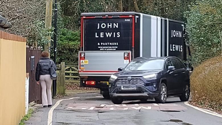 A picture of Walton Road in the village of Wavendon near Milton Keynes. You can see a woman trying to walk on a narrow path next to a road, where a John Lewis delivery van is trying to drive past a car.
