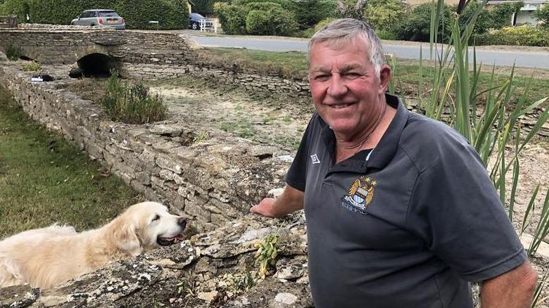 Martin Carter wearing a black polo shirt stood by the dried up River Thames in Ashton Keynes. A cream-coloured labrador dog is close by him, with a small stone wall separating them