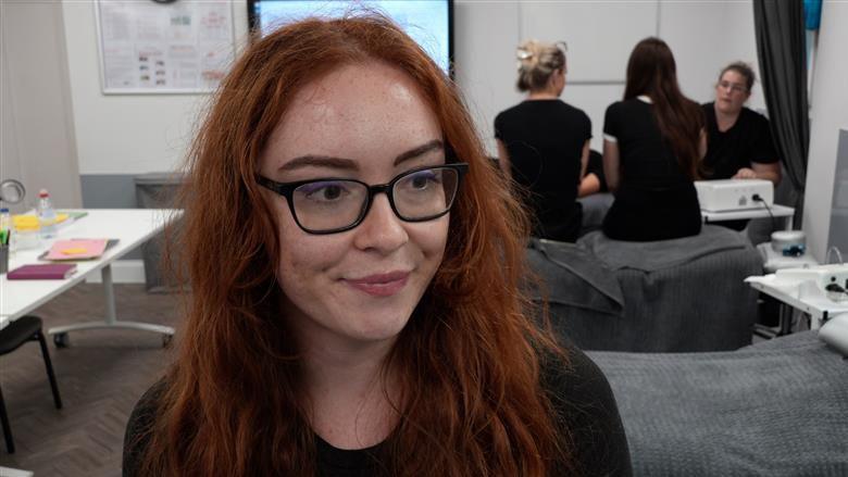 Lucy Porter is in a beauty studio, close to the camera, but looking slightly off camera to a reporter out of shot. She has red hair, freckles and glasses. She is pursing her lips slightly.