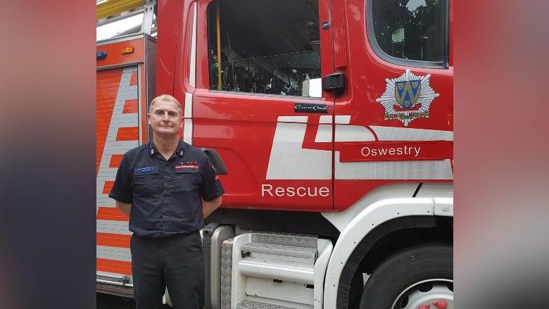 John Davies is standing in front of a red fire engine. He has his hands behind his back and is wearing a navy shirt and black trousers. The shirt has a Shropshire Fire and Rescue Service badge on the breast.