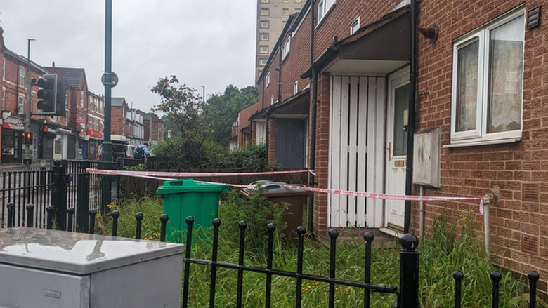 The front of the house with red and white tape around it, overgrown grass, a green and brown bin