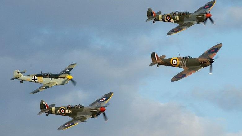 Four of the warbirds mid-flight in a cloudy sky. They are painted in shades of green, have two pilots inside, a propeller on the front and a wing at either side.