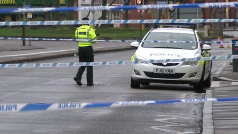 A blue and white police cordon across a road. A police officer in a high visibility jacket can be seen next to a marked police patrol car.