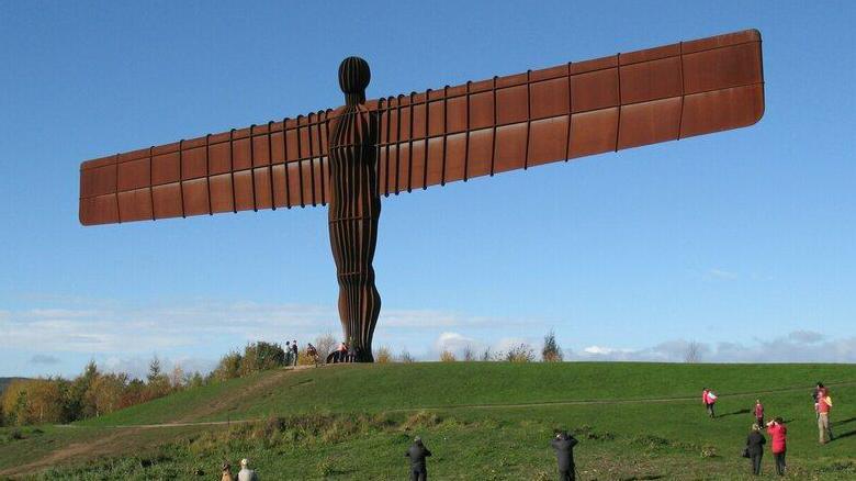 The Angel of the North, a very tall copper/brown statue with large, wide ridged wings and a ridged body. It is on a green, grassy hill, and there are some people gathered around it. The blue sky is visible in the background, with a few clouds.