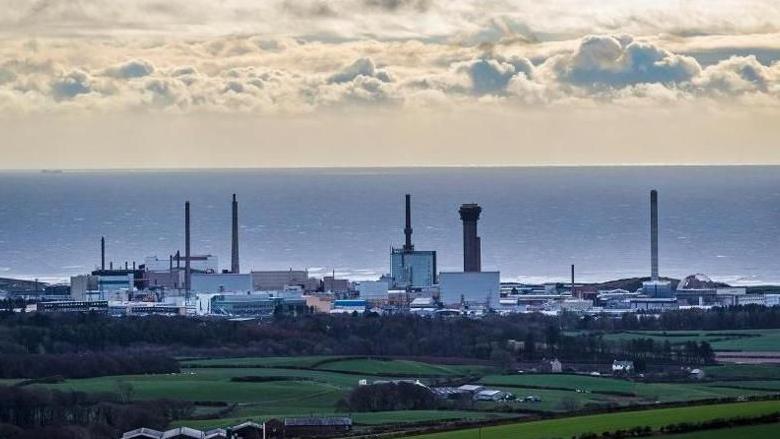 The cloudy sky, with the Sellafield nuclear site in the distance. It is made up of various large, tall buildings. Rolling fields can be seen in front.